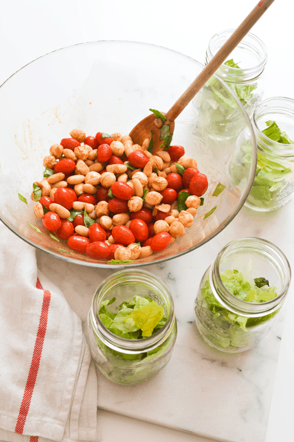 A glass bowl of white bean caprese salad on a counter next to mason jars with romaine salad in them. 