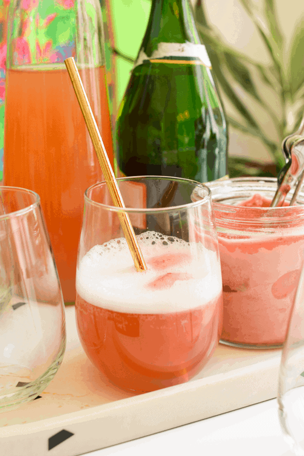 A strawberry sorbet cocktail in a stemless wine glass with a copper straw on a tray next to cocktail ingredients.