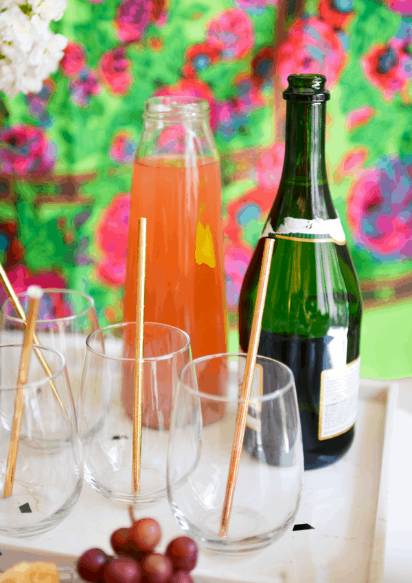 Stemless wine glasses with copper straws on a white tray with a bottle of champagne, and a bottle of strawberry lemonade.