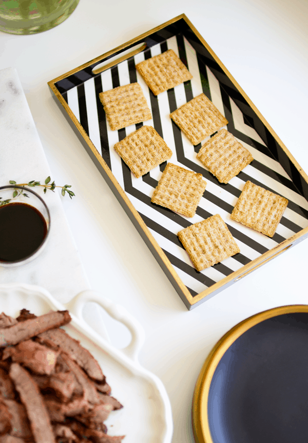A tray of Triscuits on a table next to a plate of flank steak and other ingredients to make an easy appetizer. 