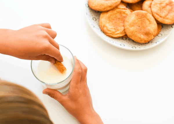 Kid dipping an Orange Snickerdoodle into a glass of milk with a plate of cookies on a table. 