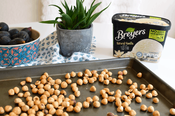 Blanched and toasted hazel nuts on a baking sheet in front of a carton of ice cream and a bowl full of figs.