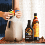 Woman arranging straws in a milk jug full of a white russian recipe next to a bottle of Kahlua and chocolate syrup.