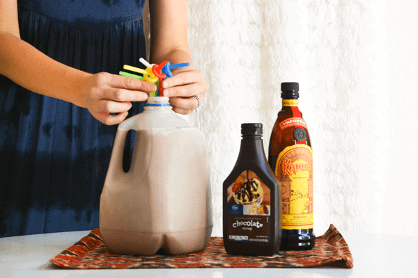 Woman arranging straws in a milk jug full of a white russian recipe next to a bottle of Kahlua and chocolate syrup.