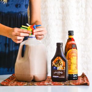 Woman arranging straws in a milk jug full of large batch white Russian, next to Kahlua and chocolate syrup.