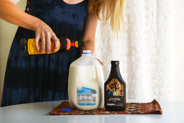 Woman pouring Kahlua into a milk jug, next to a bottle of chocolate syrup.