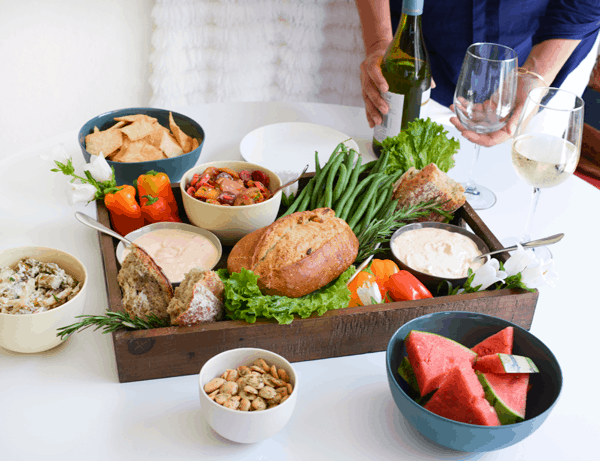 A snack board on a table with a woman holding wine behind it. 