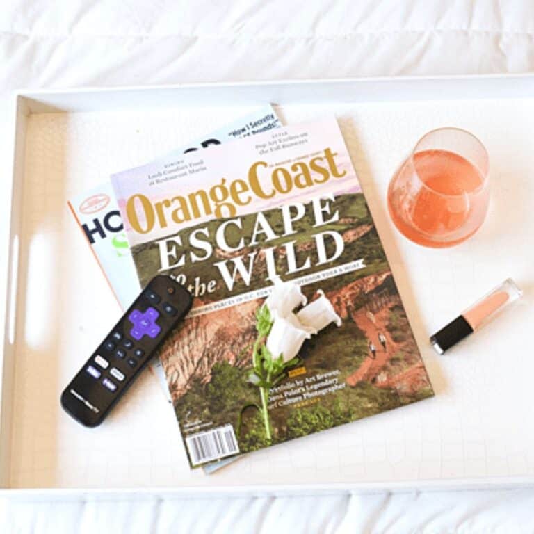 A close up of some magazines on a white bed tray with a TV remote, nail polish and a glass of rose.