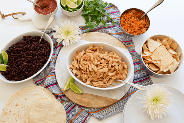 Overhead view of serving bowls with shredded chicken, black beans and other toppings for tacos. 