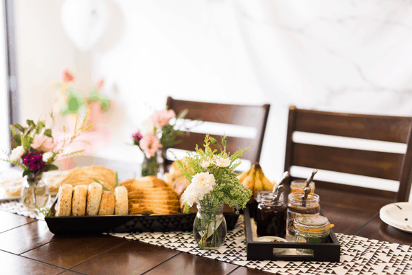 A brunch table holding fresh flowers and trays of bagels and breads.
