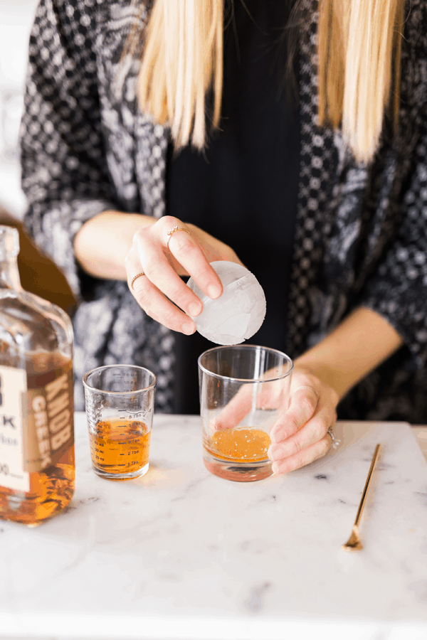 Woman adding a big round ice cube to a short cocktail glass.