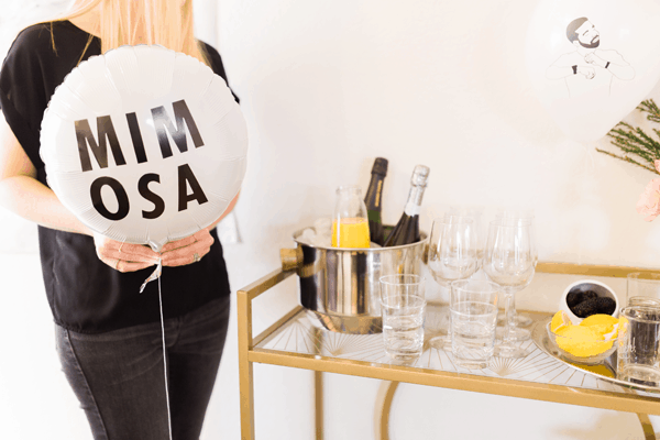 Woman holding a balloon that says MIMOSA standing next to a bar cart with mimosa ingredients.