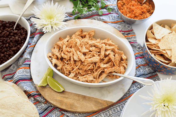 Close up of a white bowl with seasoned shredded chicken next to a bowl of chips and black beans. 