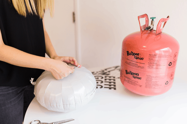 Woman adding letter stickers to a blown up white mylar balloon on a table next to a helium tank. 