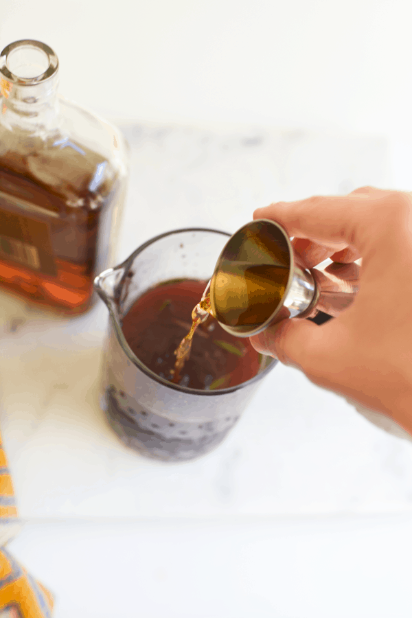 A woman pouring whiskey into a bar pitcher holding the rest of the ingredients for a Blackberry Whiskey Lemonade.
