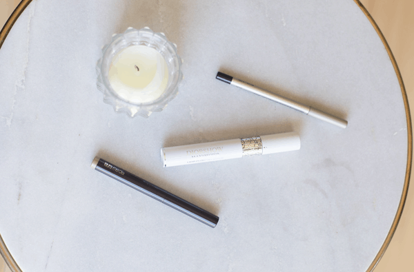 An eye shadow stick, and eyeliner and a mascara on a table next to a candle. 