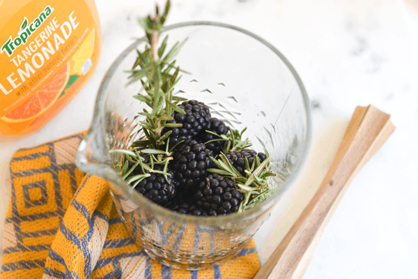 A bar pitcher with fresh blackberries and rosemary next to a bottle of lemonade and a wooden muddler.
