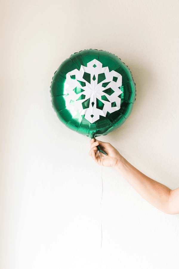 A woman's hand holding a green mylar balloon with a snowflake on it in front of a wall. 