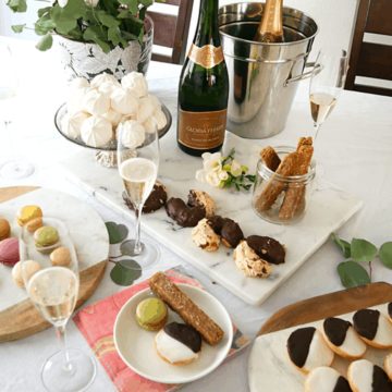 Close up of plates of cookies on a table next to a few bottles of champagne and flowers.