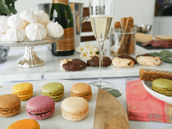Colorful macaron cookies on a tray with champagne next to it on a table surrounded by other plates of cookies.