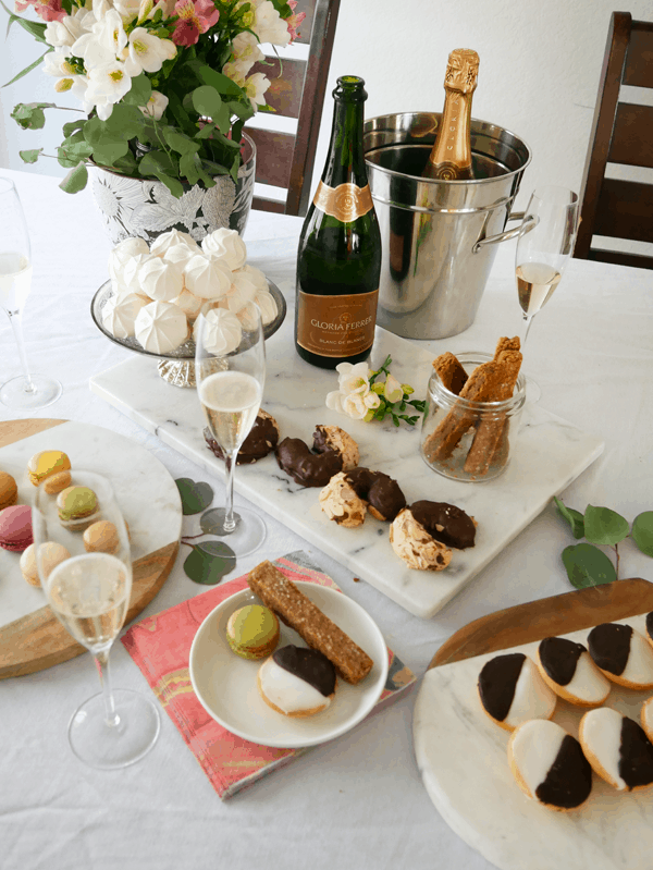 Plates of cookies on a table next to a bottle of champagne and champagne flutes for a cookie exchange party.