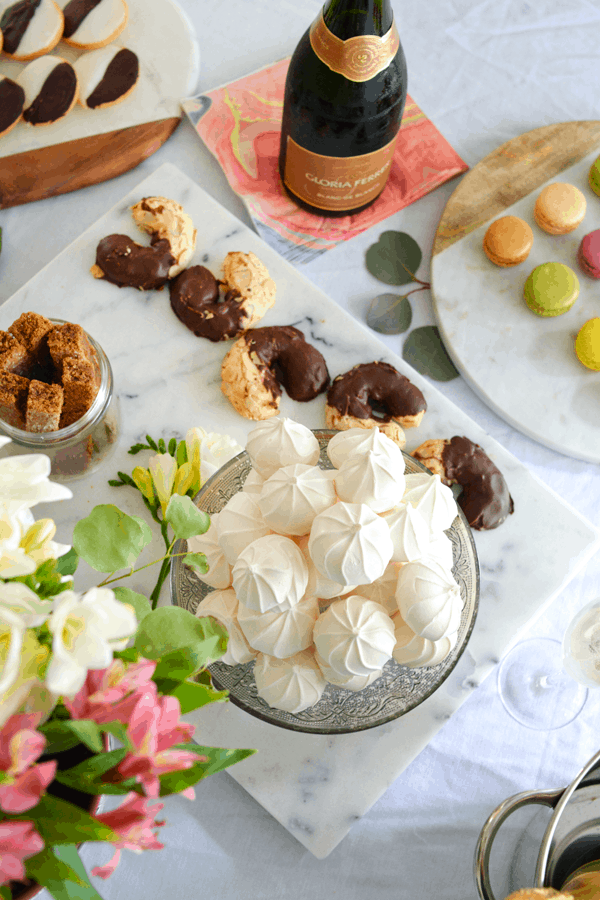 An overhead shot of a plate of white meringue cookies next to flowers, other plates of cookies and a bottle of champagne.