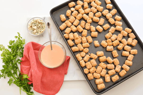 A tray of tater tots next to a bowl of Buffalo aioli and a smaller bowl of blue cheese crumbles. 