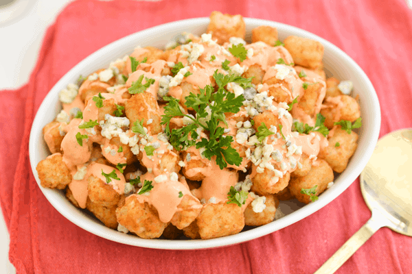 Loaded tater tots with blue cheese and spicy aioli in a white baking dish on a red napkin.  