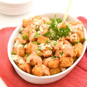 Tater tots in a while baking dish drizzled with Buffalo aioli, blue cheese and parsley.