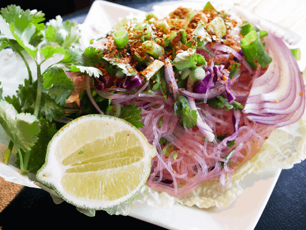 A salad of fresh vegetables on top of a bed of purple glass noodles next to a half of a lime and cilantro.