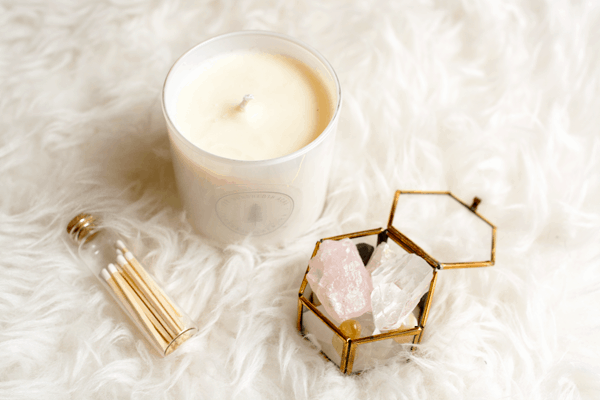 Image contains a candle, matches and crystals in a glass container on a furry white rug.