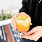 Peach paloma recipe in clear cocktail glass held by a woman in black shirt.