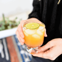 Peach paloma recipe in clear cocktail glass held by a woman in black shirt.