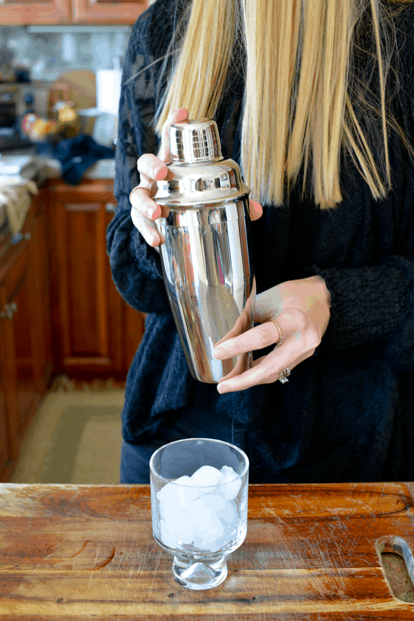 Blonde woman shaking a silver cocktail shaker filled with a peach paloma cocktail. 