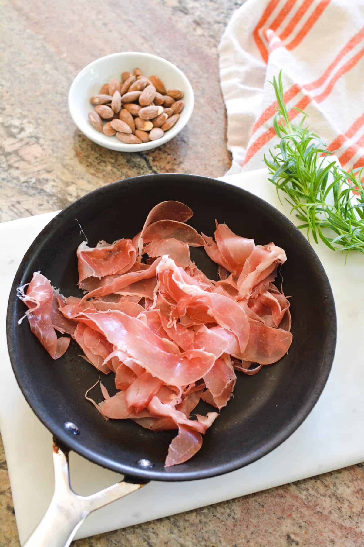 Prosciutto in a saute pan on the counter next to a bowl of almonds and fresh rosemary.