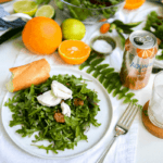 A plate holding an arugula salad topped with pecans, beets and goat cheese on a table next to a canned drink with fresh citrus in the background.