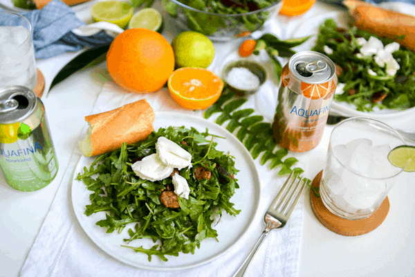 A plate holding an arugula salad topped with pecans, beets and goat cheese on a table next to a canned drink with fresh citrus in the background.