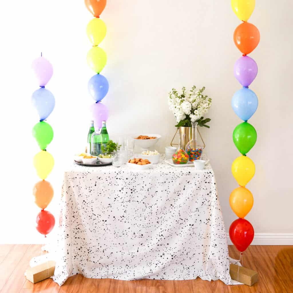 A party table with 3 rainbow balloon columns next to it.