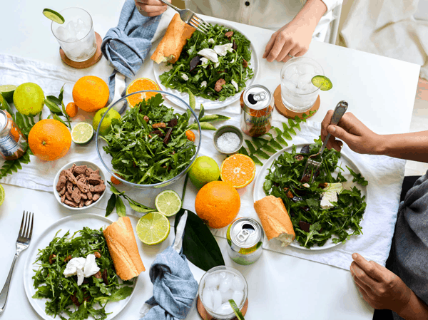 Overhead shot of a lunch table with arugula salad, fresh citrus and canned drinks.
