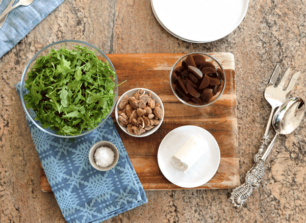 A bowl of arugula, goat cheese, salt, beets and candied pecans on a cutting board.