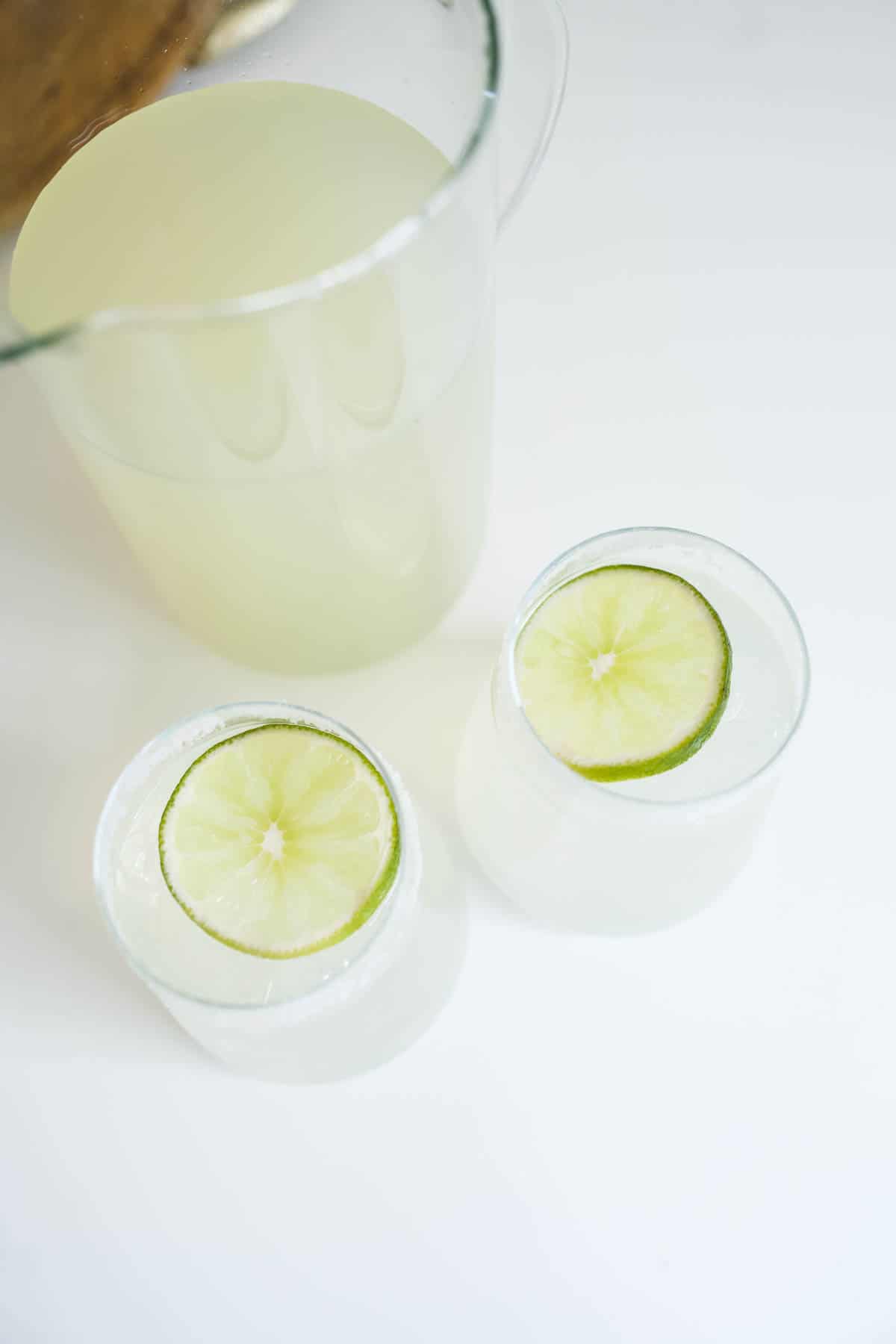 Two salt rimmed glasses holding margarita and lime garnish next to a pitcher of margaritas on a table.