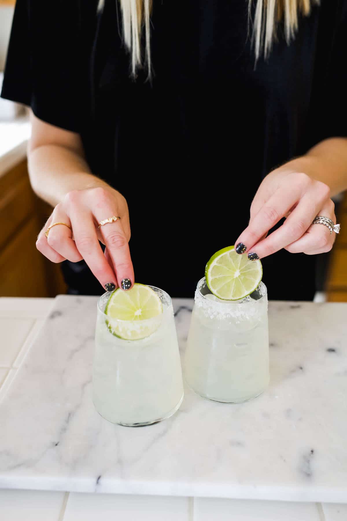 Woman adding lime wheels to two glasses full of margaritas on a counter.