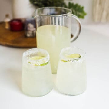 2 salt rimmed glasses holding margarita drinks next to a pitcher of margaritas on a table.