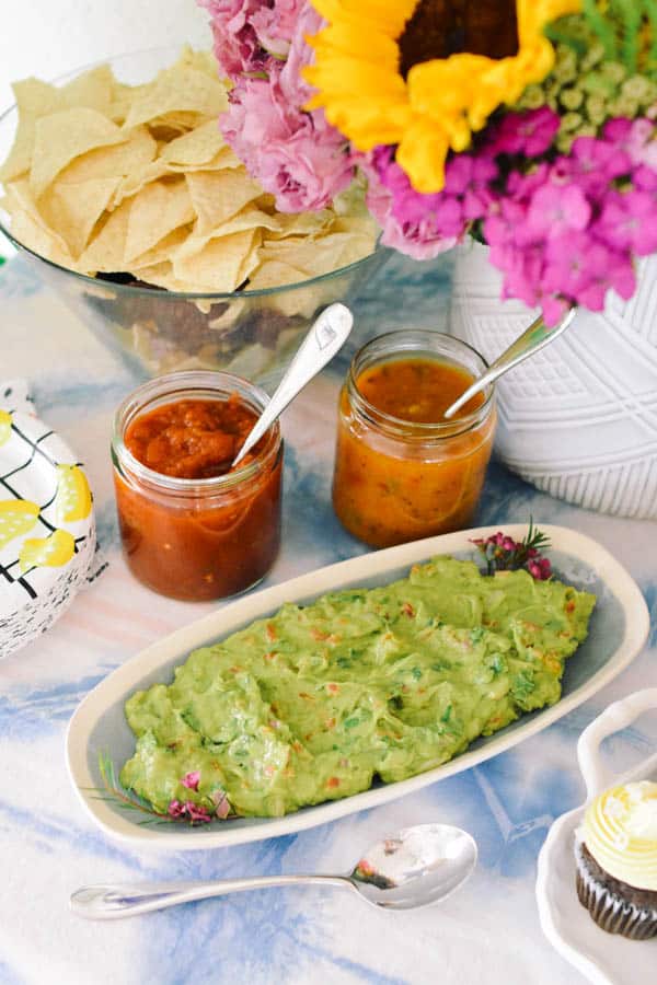 A tray of guacamole sitting on a food table next to a bowl of chips and a few types of salsa for an easy graduation party
