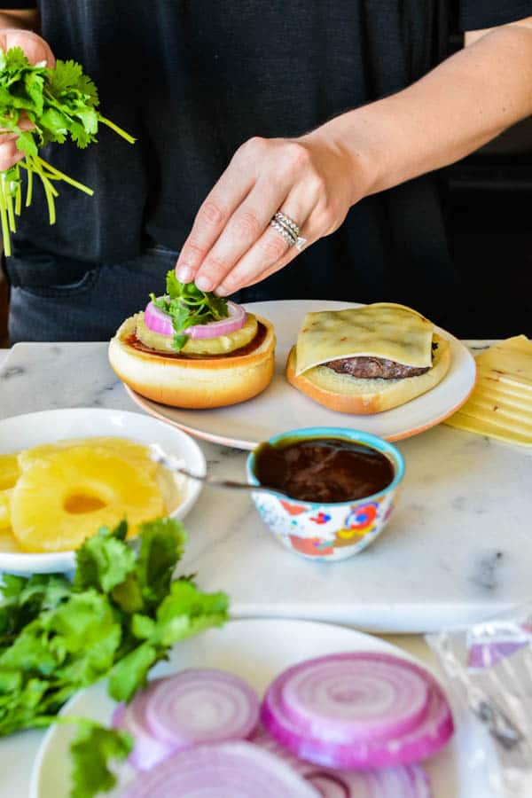 Woman adding cilantro to the top of a  hamburger with pineapple, and red onion. 