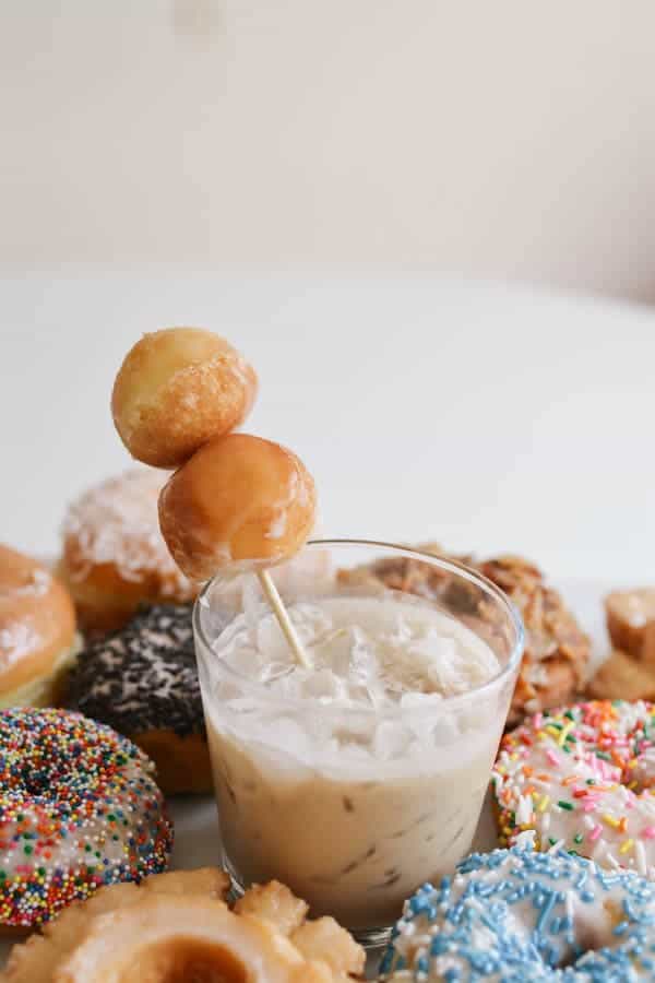 A Coffee White Russian cocktail in a glass with a skewer with donut holes surrounded by donuts on the table. 