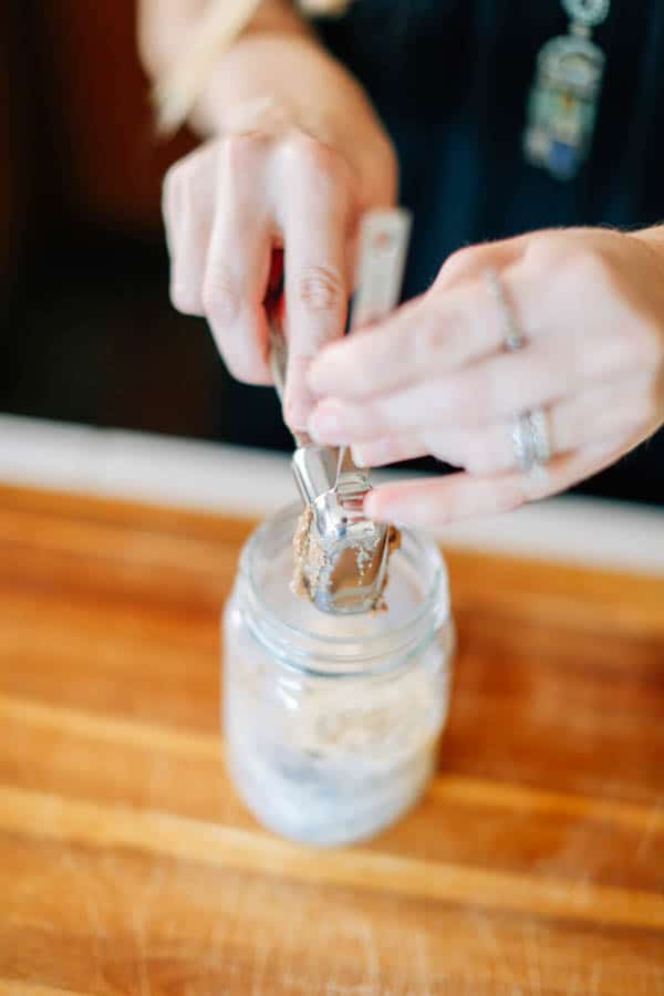 A woman using measuring spoons to add almond butter to a mason jar with rolled oats.