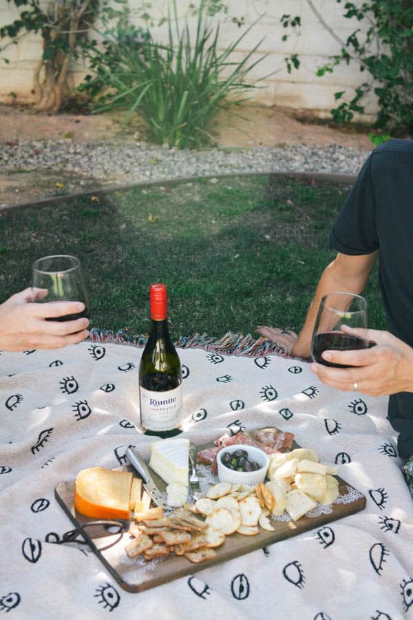 A man and woman enjoying a charcuterie board on a picnic blanket and holding wine glasses.