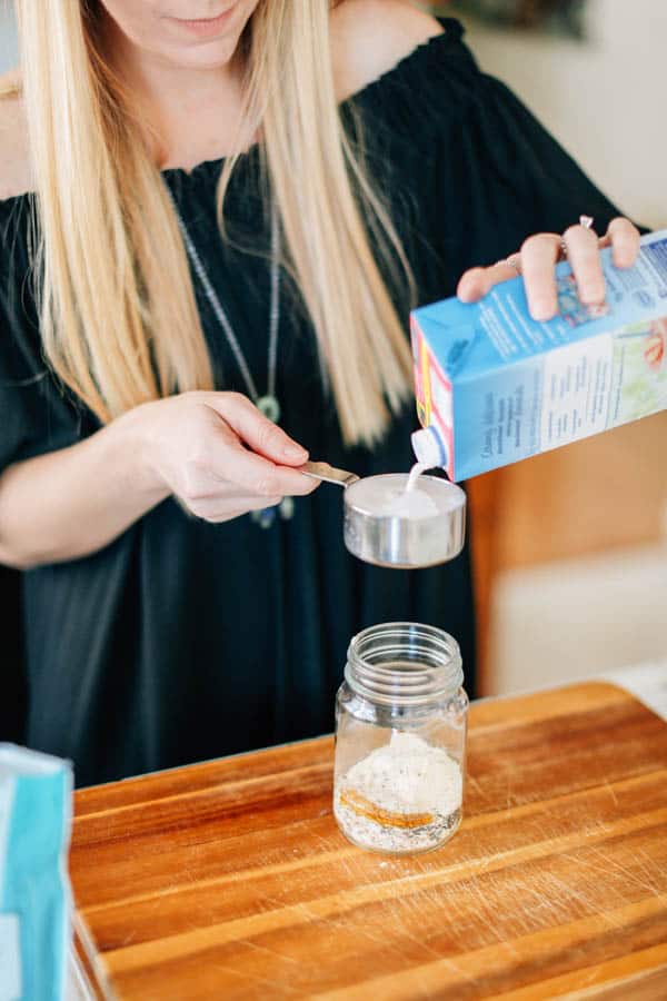 A woman pouring almond milk into a measuring cup over a jar of dry overnight oats ingredients.