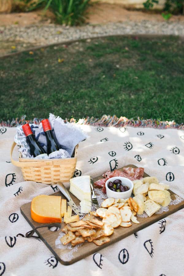 A charcuterie board picnic on a blanket on grass with a basket and small wine bottles.